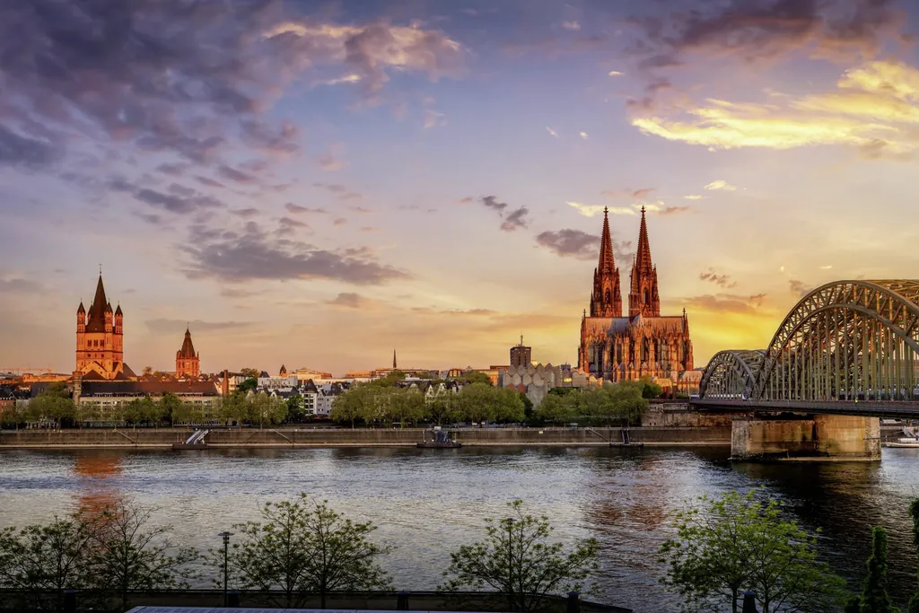 Blick auf den Kölner Dom und die Hohenzollernbrücke bei Sonnenuntergang, mit Rhein und Altstadt im Vordergrund