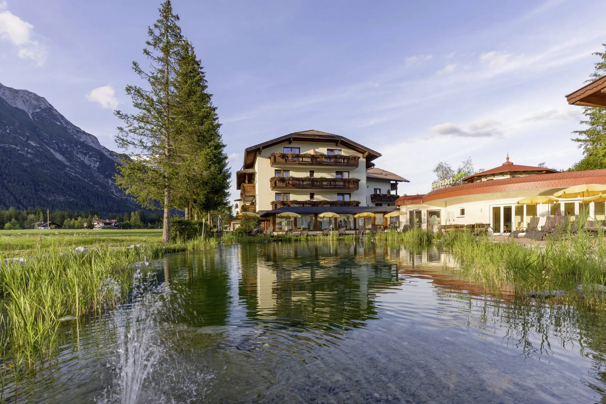Blick über den Naturteich des Alpenhotel Karwendel umringt von Bäumen auf die Fassade mit Holzbalkonen