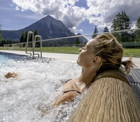 blonde Frau mit Zopf liegt am Rand der Whirlliege im Outdoorpool des Alpenhotel Karwendel