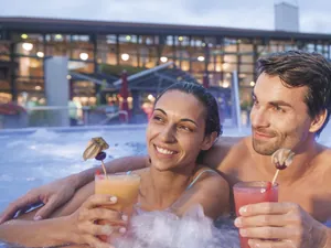 Entspanntes Paar im Outdoorpool der Obermain Therme, hält farbige Cocktails, beleuchtetes Gebäude im Hintergrund
