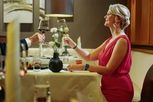 Frau in rotem Kleid bei einem romantischen Dinner, hält ein Glas Rotwein zum Anstoßen, dekorativer Tisch mit Blumenvase im Best Western Plus Kurhotel an der Obermaintherme