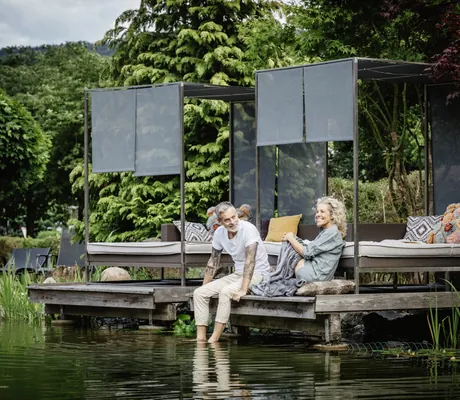 blonde Frau und grauhaariger Mann sitzen auf der Holzinsel am Naturteich im Bodenmaiser Hof