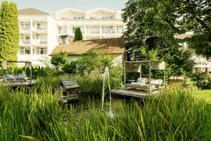 Blick auf den Naturteich des Das Parkhotel mit Springbrunnen, von Pflanzen umgebenen Daybeds und der weißen Fassade des Wellnesshotels im Hintergrund