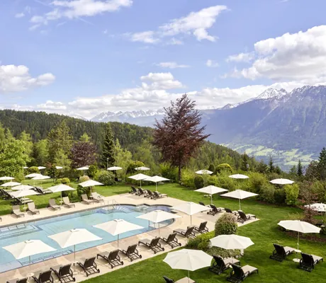 Outdoorpool des Interalpen-Hotel Tyrol mit Liegen und Sonnenschirmen, eingebettet in eine grüne Berglandschaft mit Alpenpanorama