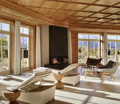 Eleganter Ruheraum im Interalpen-Hotel Tyrol mit Liegen und Kamin, umgeben von großen Fensterfronten mit Blick auf den Garten und die Berge
