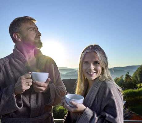 blonde Frau und Mann mit Bart stehen in Bademänteln auf dem Balkon im Wellnesshotel Zum Bräu und trinken Kaffee
