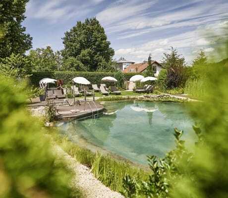 Blick auf den Naturbadeteich des Wellnesshotel Zum Bräu mit weißen Sonnenschirmen und beigen Sonnenliegen