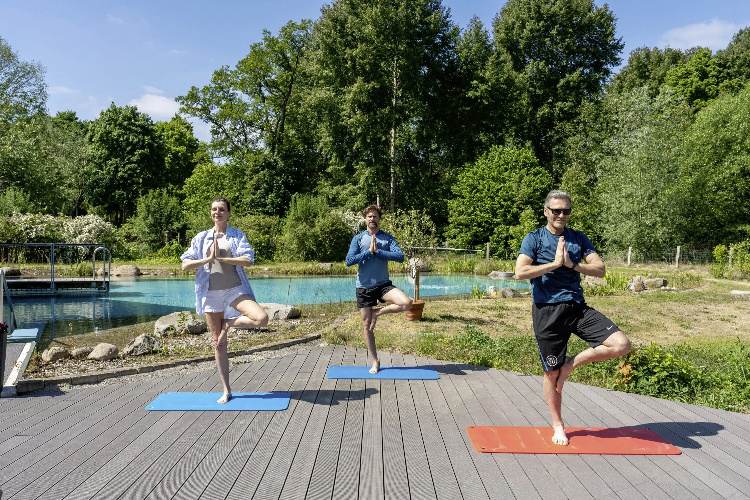 Zwei Männer und eine Frau bei einer stehenden Yogaübung auf der Sonnenterrasse des BEECH Resort Plauer See
