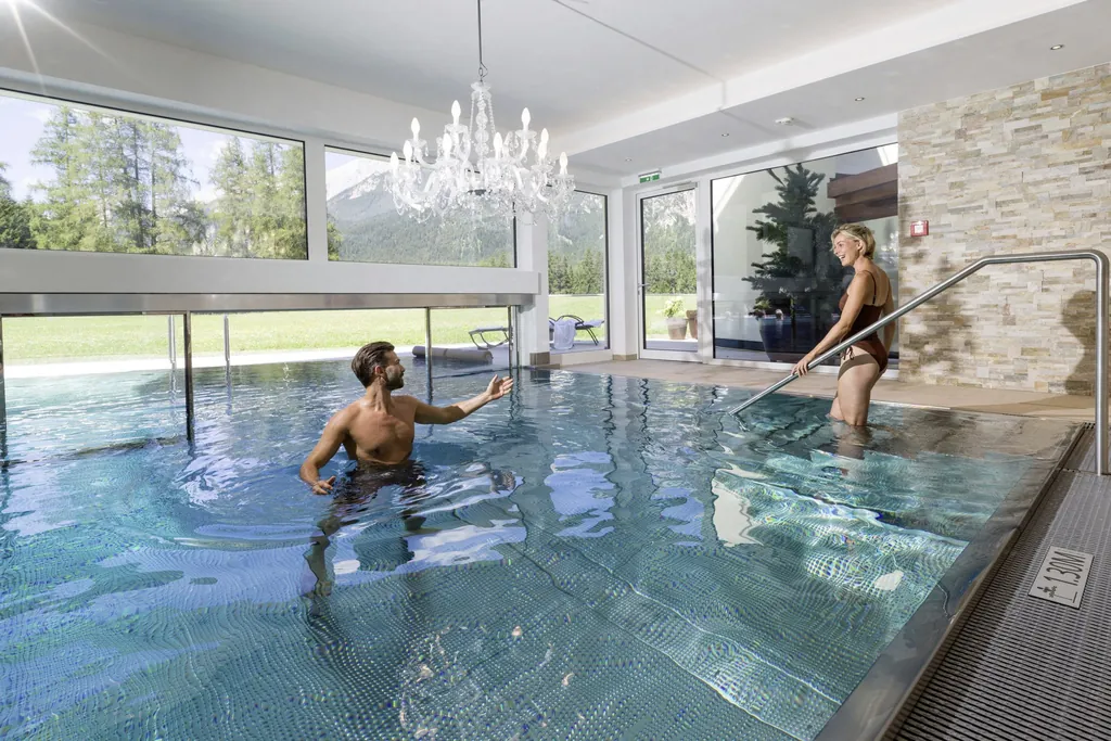 Mann steht im Indoorpool des Alpenhotel Karwendel mit Kronleuchter und winkt seiner Frau am Einstieg zu