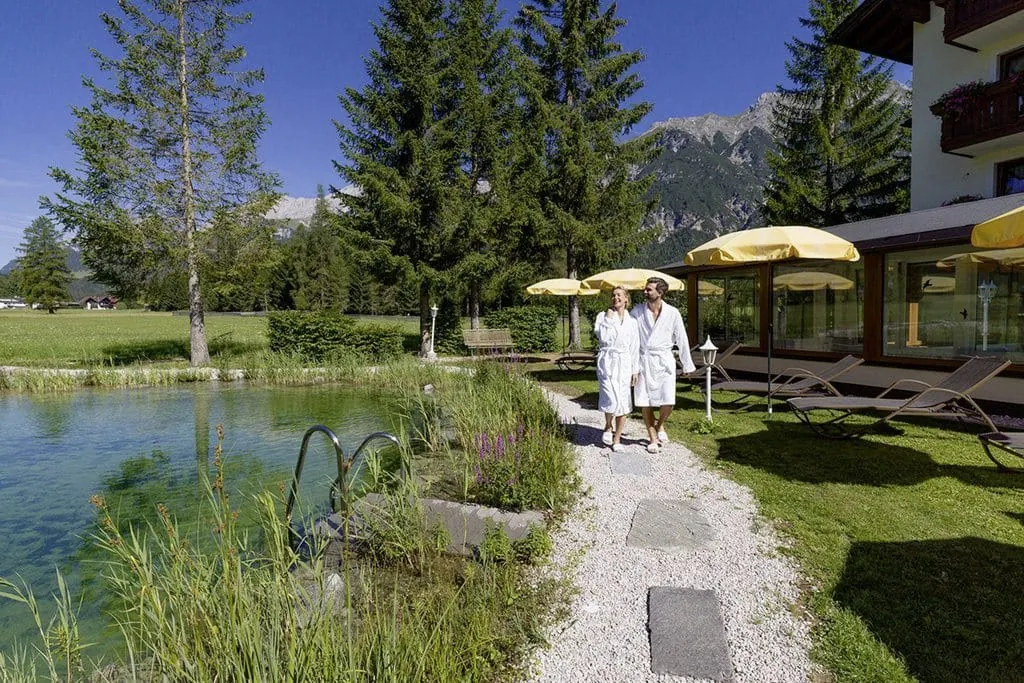 Paar in weißem Bademantel spaziert am Naturschwimmteich des Alpenhotel Karwendel entlang