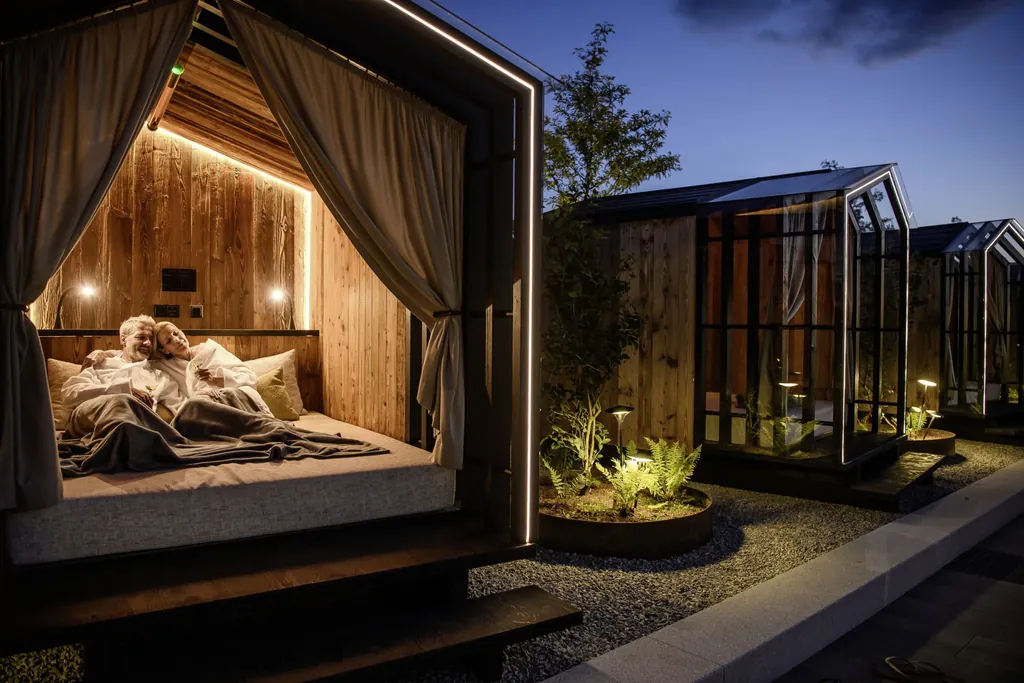 Paar in einem beleuchteten, h&ouml;lzernen Daybed mit Vorh&auml;ngen und gem&uuml;tlicher Einrichtung im DAS STEMP bei Abendd&auml;mmerung
