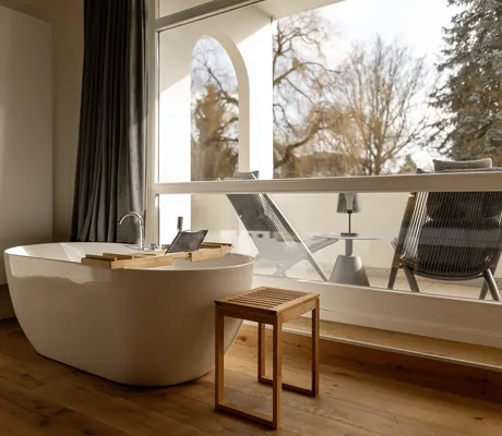 Freistehende Badewanne mit Holzablage vor bodentiefem Fenster mit Blick auf eine Terrasse mit bequemen Stühlen im Das Parkhotel