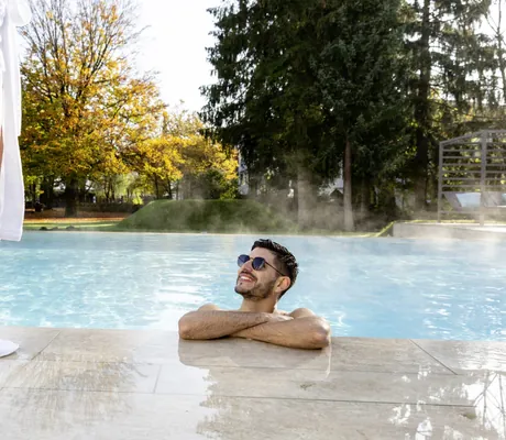 Mann im beheizten Outdoorpool des Das Parkhotel mit Blick auf herbstliche Bäume, während eine Person mit Bademantel am Beckenrand steht