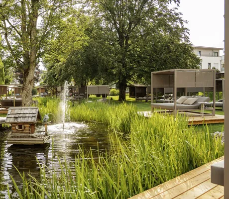 Naturteich im Wellnesshotel Das Parkhotel mit schwimmender Holzdekoration, Wasserfontäne, umgebendem Schilf und Daybeds auf Holzplattformen