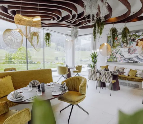Stilvolles Restaurant im Naturresort PURADIES mit geschwungenen Decken, Wandkunst, hängenden Pflanzen und großen Fenstern mit Blick ins Grüne