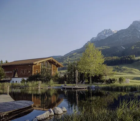 Naturbadeteich des Naturresort PURADIES mit Holzstegen, umgeben von grüner Landschaft und Bergpanorama im Hintergrund