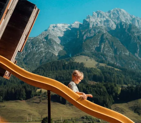 Junge auf orangefarbener Rutsche des Abenteuerspielplatzes im Naturresort PURADIES mit malerischem Blick auf die Leoganger Berge