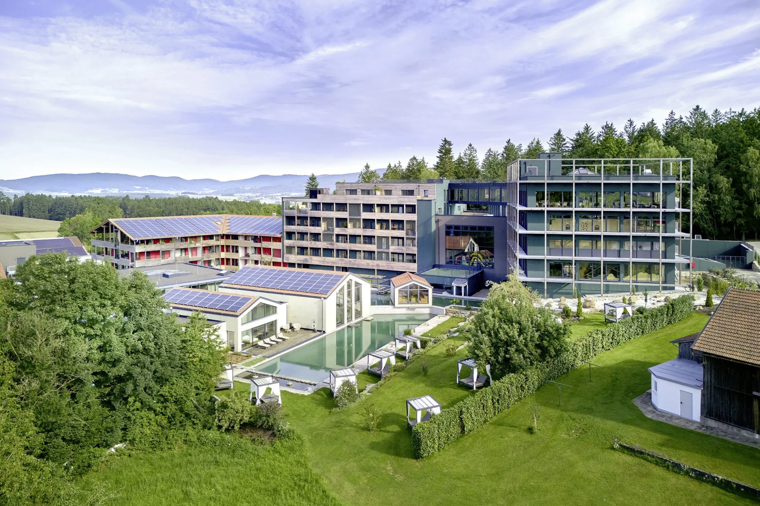 Außenansicht des ULRICHSHOF – Nature · Family · Design mit moderner Architektur, Solarzellen auf den Dächern, Naturpool und umliegenden Liegeflächen, eingebettet in eine grüne Landschaft mit Wald und Hügeln im Hintergrund
