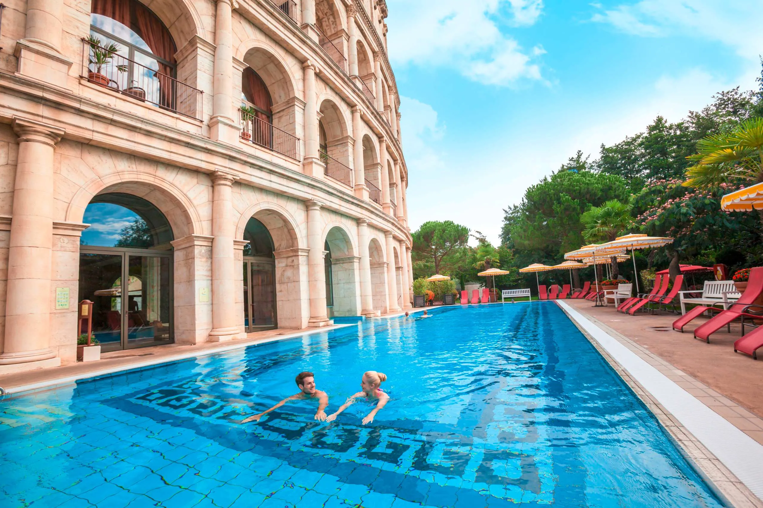 Outdoorpool des Erlebnishotels Colosseo Europa-Park in Rust mit römischer Fassadenarchitektur, roten Liegestühlen und zwei schwimmenden Personen