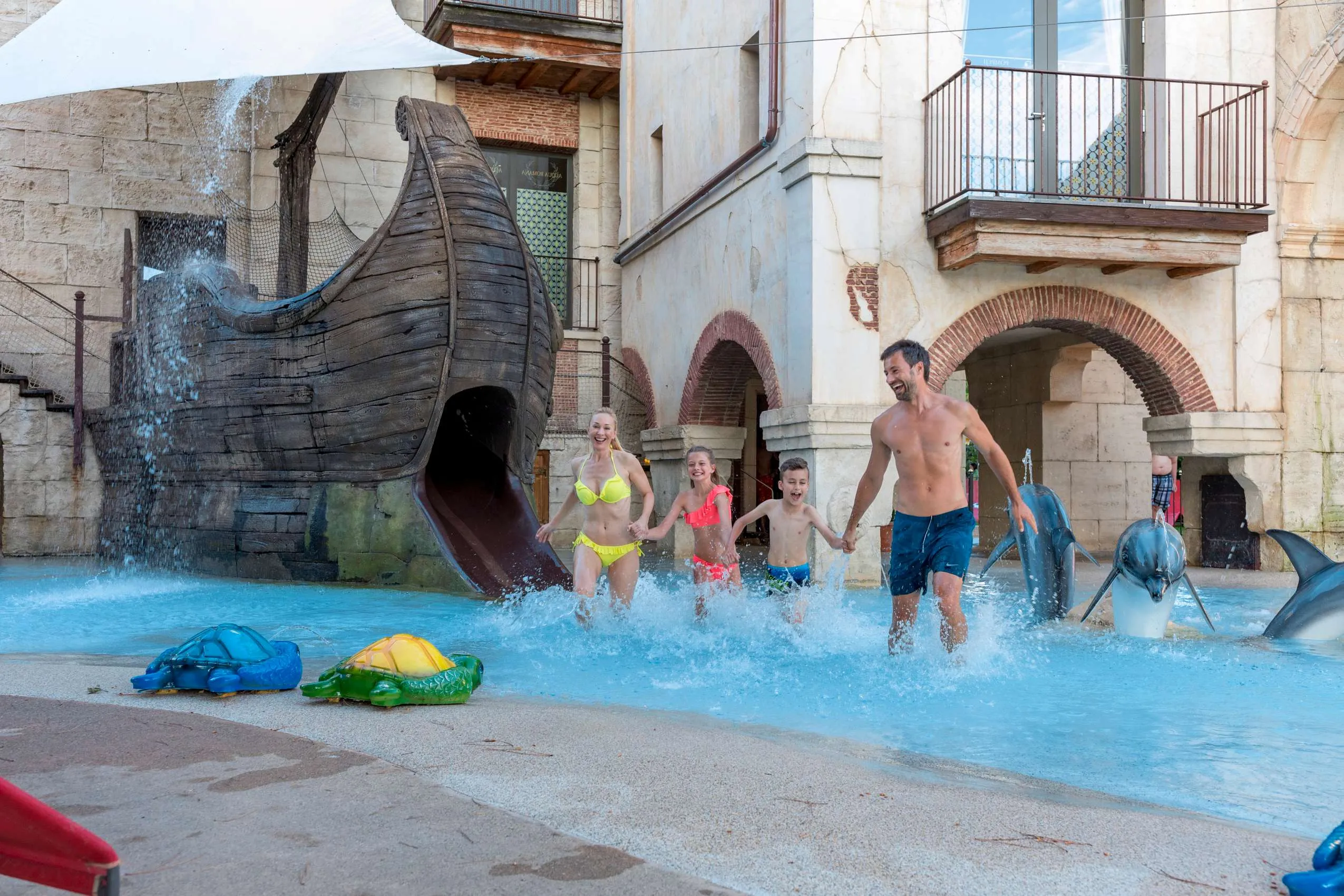Familie im Wasserspielbereich des Erlebnishotels Colosseo Europa-Park in Rust mit Holzboot-Rutsche, Delfin-Skulpturen und spritzendem Wasser