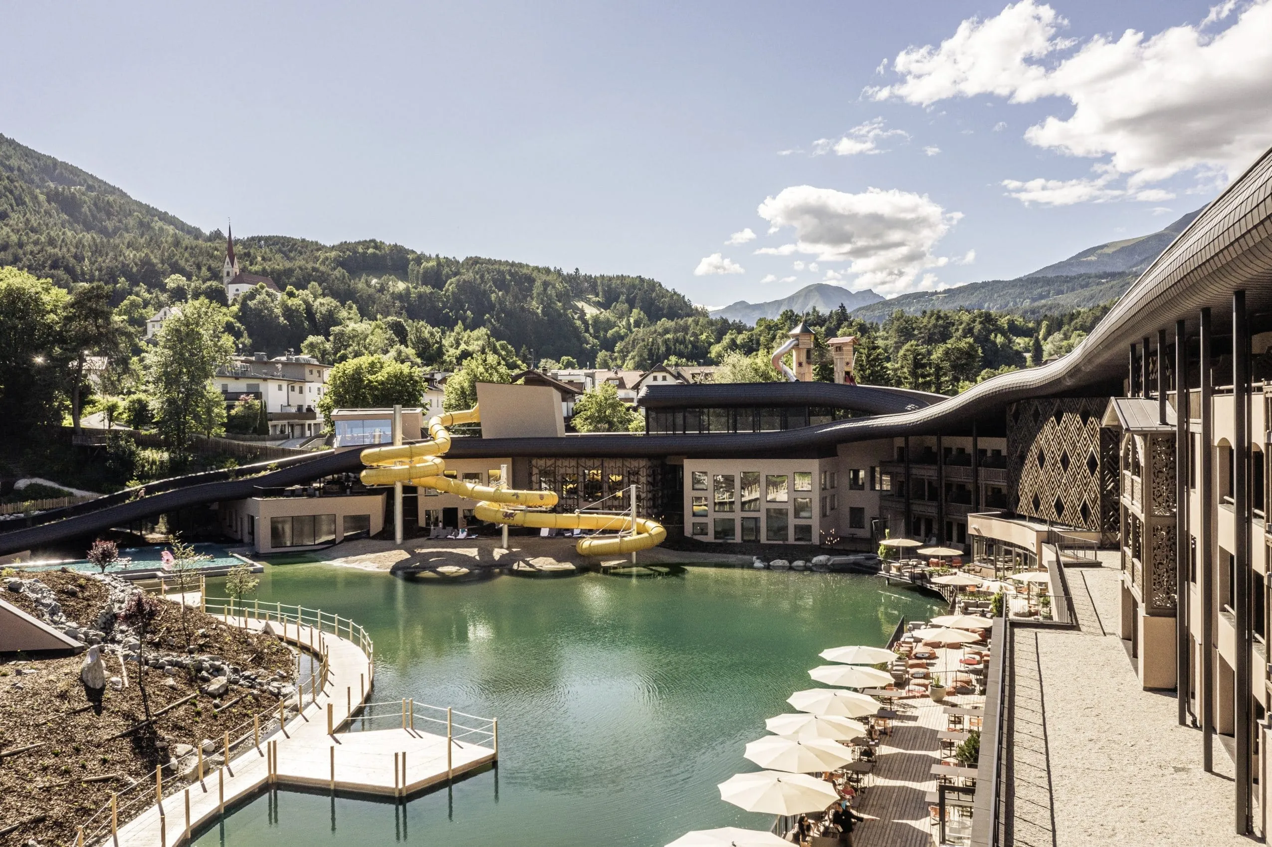 Hotelanlage des Falkensteiner Family Resort Lido von außen mit See, Wasserrutsche, Terrasse und Ausblick auf Wald und Berge