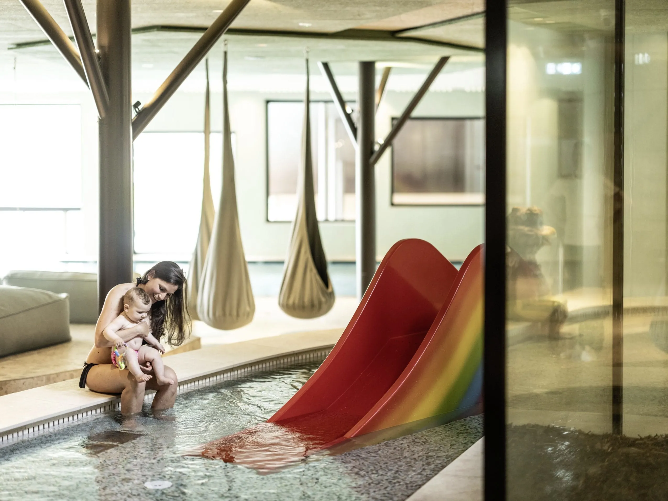 Junge Frau sitzt mit Baby auf dem Schoß am Rand des Babybeckens mit kleiner Wasserrutsche im Falkensteiner Family Resort Lido