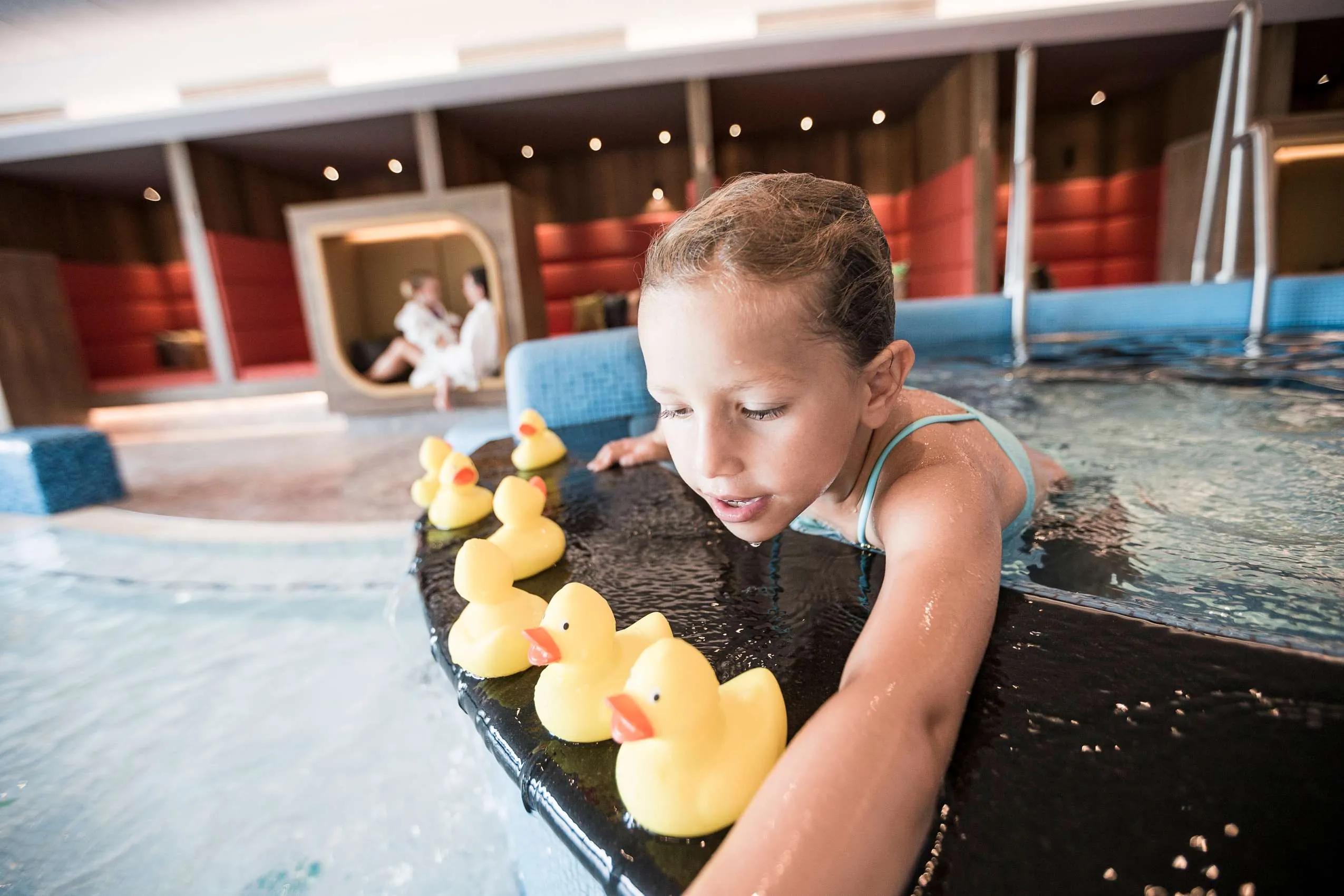 Kind im Kinderbecken des Familotel Allgäuer Berghof, spielend mit gelben Plastikenten, im Hintergrund gemütliche Sitzbereiche