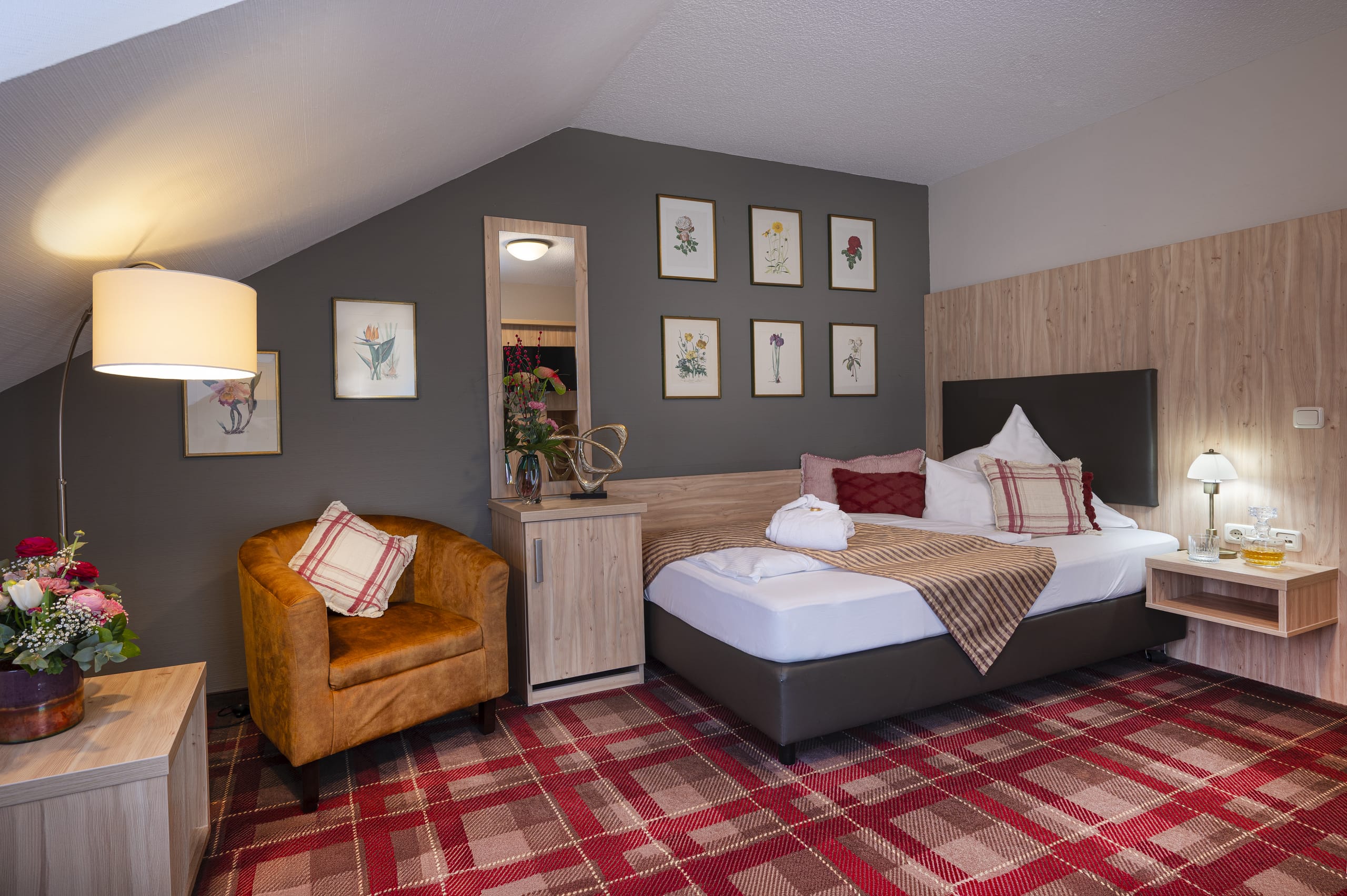 Gemütlich eingerichtetes Einzelzimmer im Tagungshotel Ringhotel Sonneck mit floralen Kunstwerken, rotem Teppich und einem bequemen Sessel.