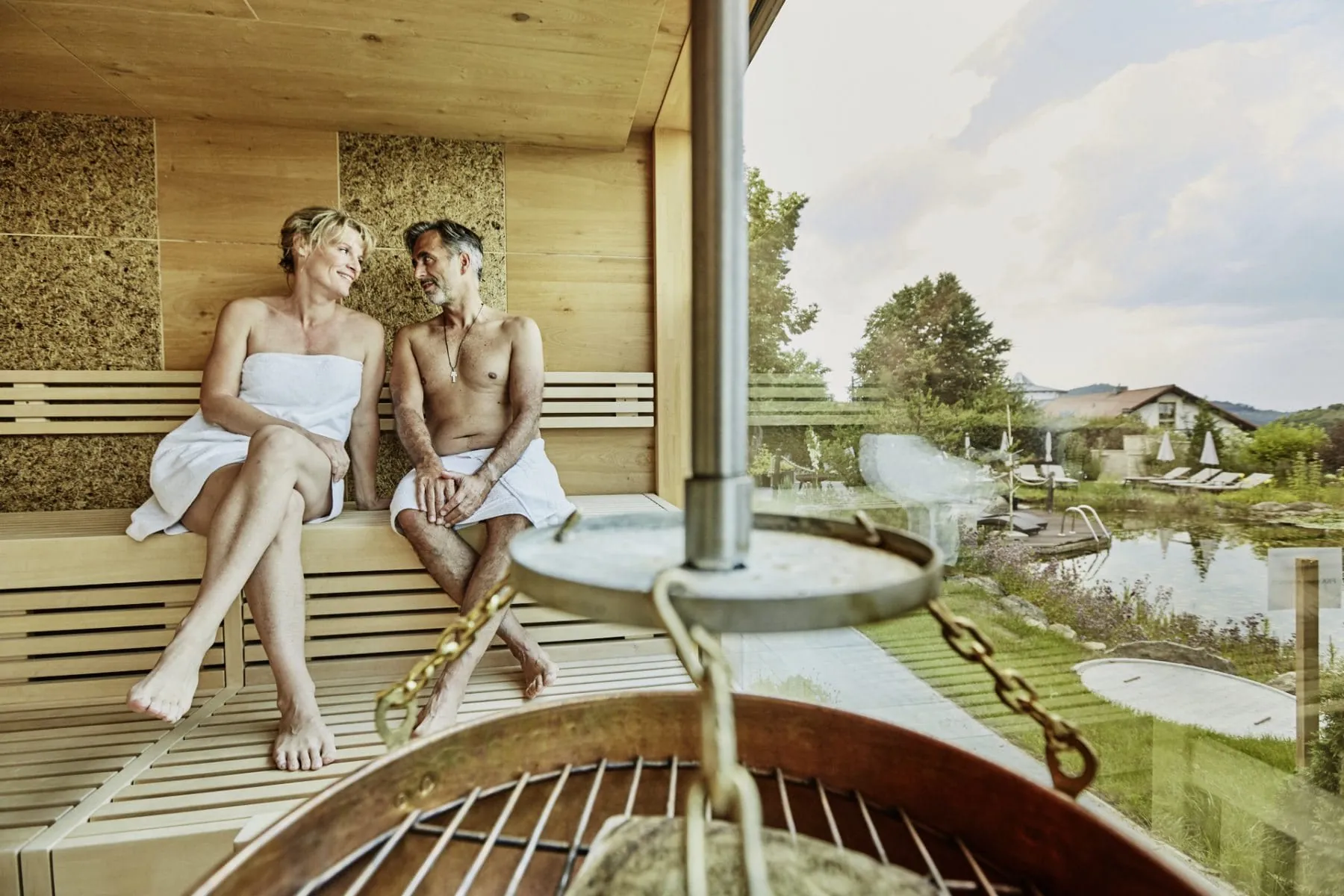 Paar mit wei&szlig;en Handt&uuml;chern sitzt in der Sauna des Wellnesshotel Zum Br&auml;u mit Blick auf den Naturbadeteich